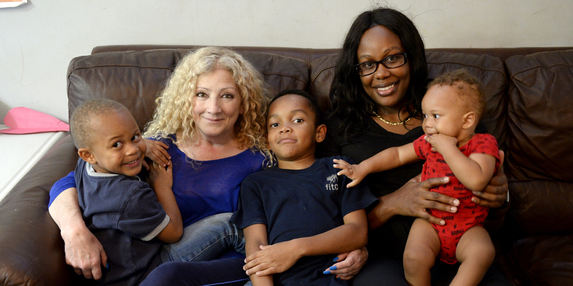 A mom and her three young children sit on the couch next to another woman who's providing home visitation services to them.