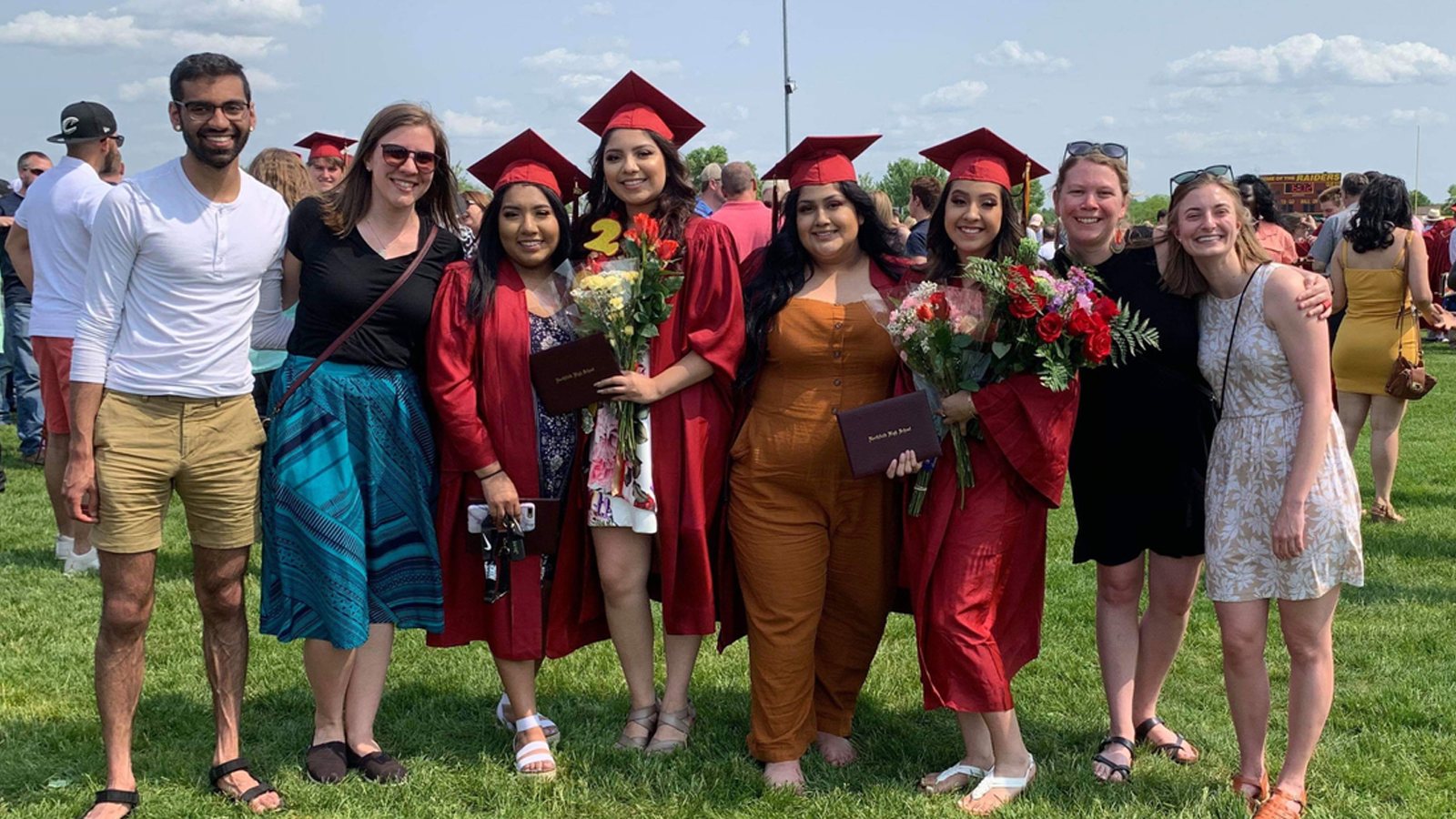 A group of high school graduates wearing caps and gowns smile at the camera with their family and friends.