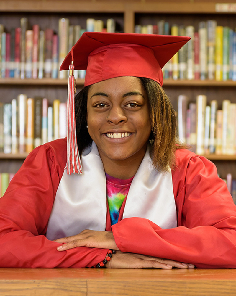 A high school graduate smiles at the camera. She's wearing a red cap and gown.
