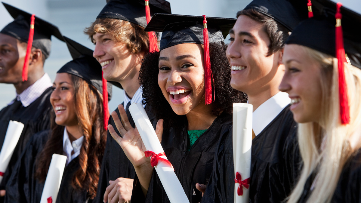A group of high school graduates stand in a line. The photo focuses on one young woman waving and smiling at the camera.