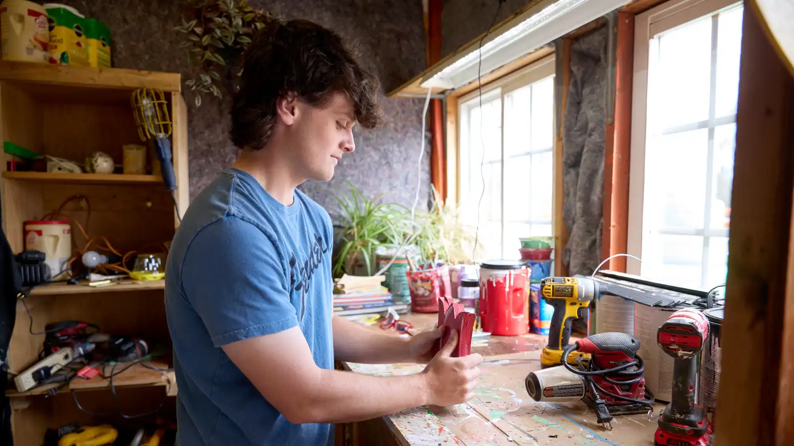 A high school-aged boy stands at a table in a workshop, glueing two items together.