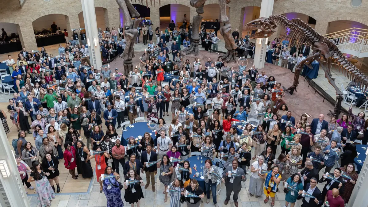 A large group of Cradle to Career Network members smile up at the camera at the 2025 Cradle to Career Network convening. They're in a museum and standing below a large mounted skeleton of a dinosaur