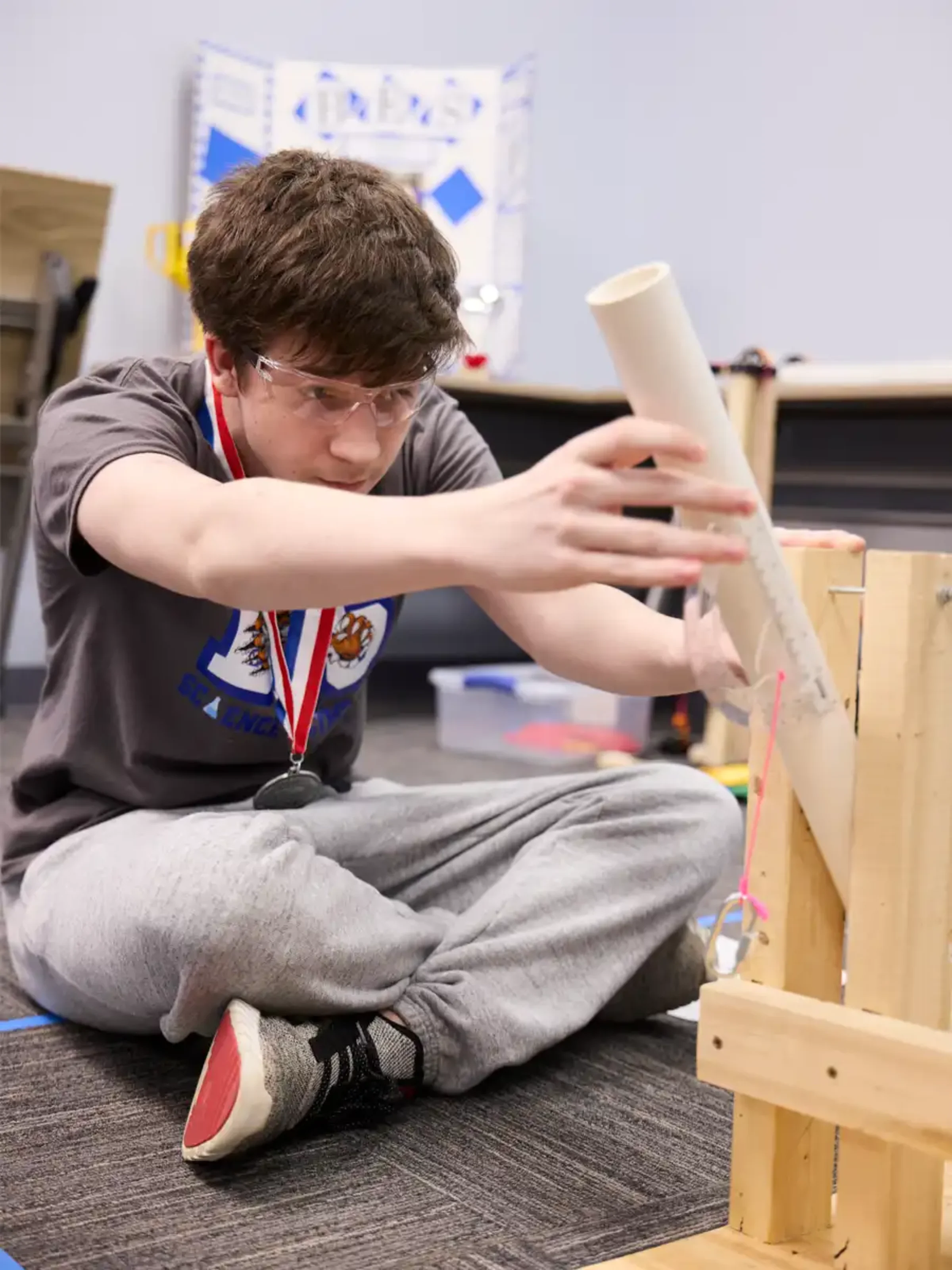 A high school boy is sitting on the ground wearing protective goggles and building a structure out of wood.