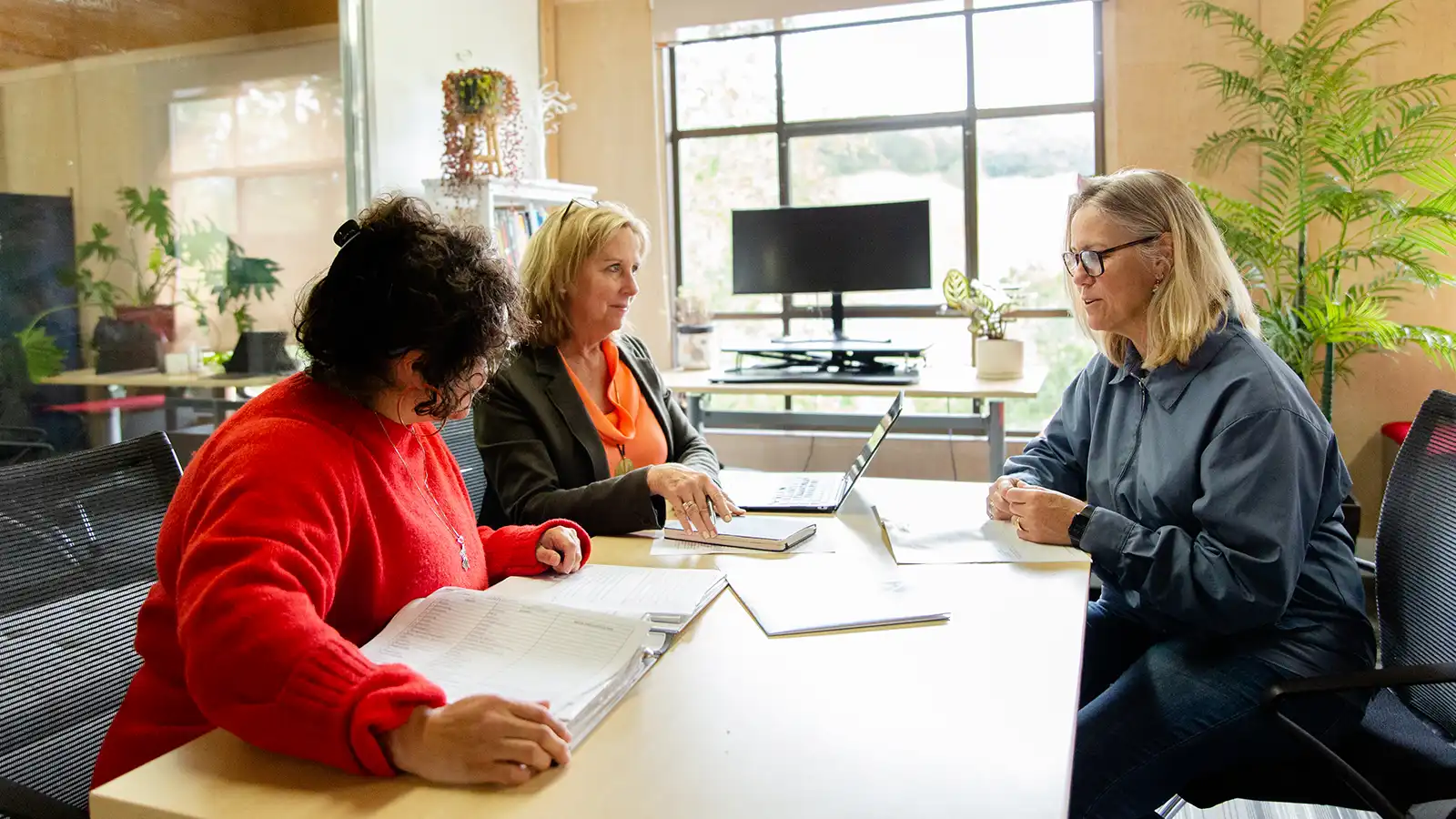 Three women sit at a table in an office space having a discussion.