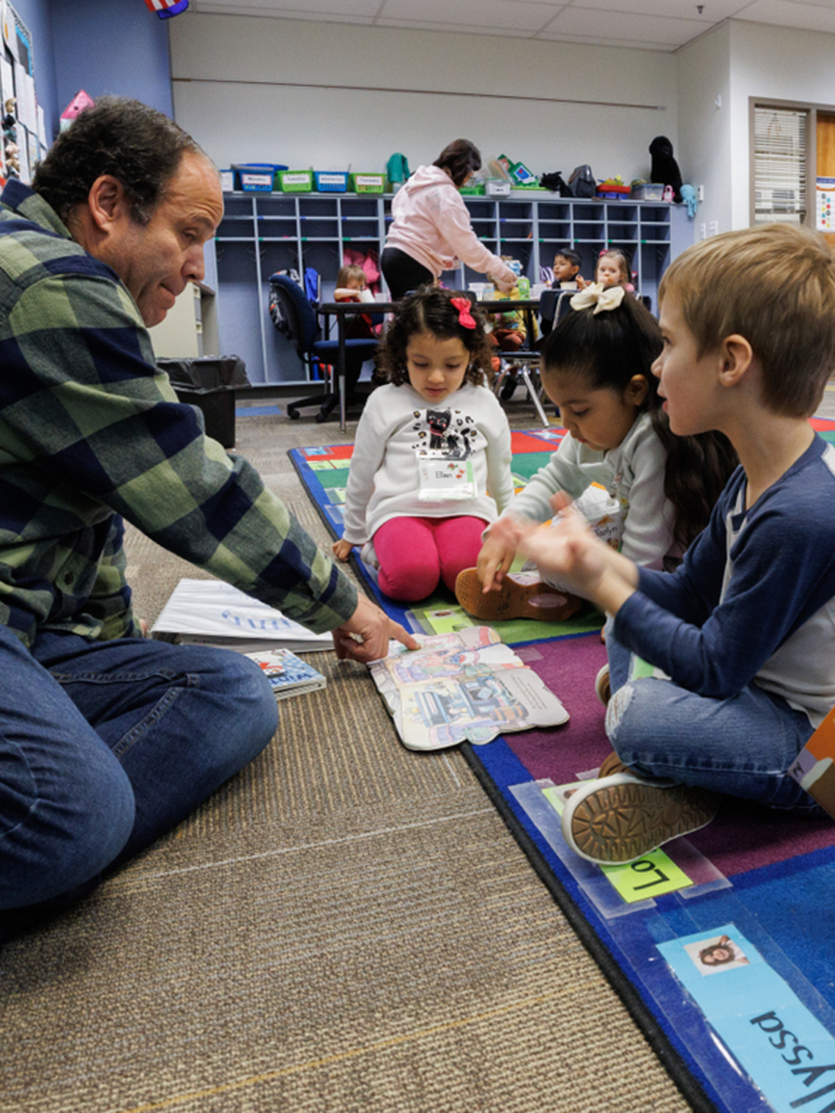 A teacher sits on the ground in front of three young children, pointing at a book laying open in front of them.