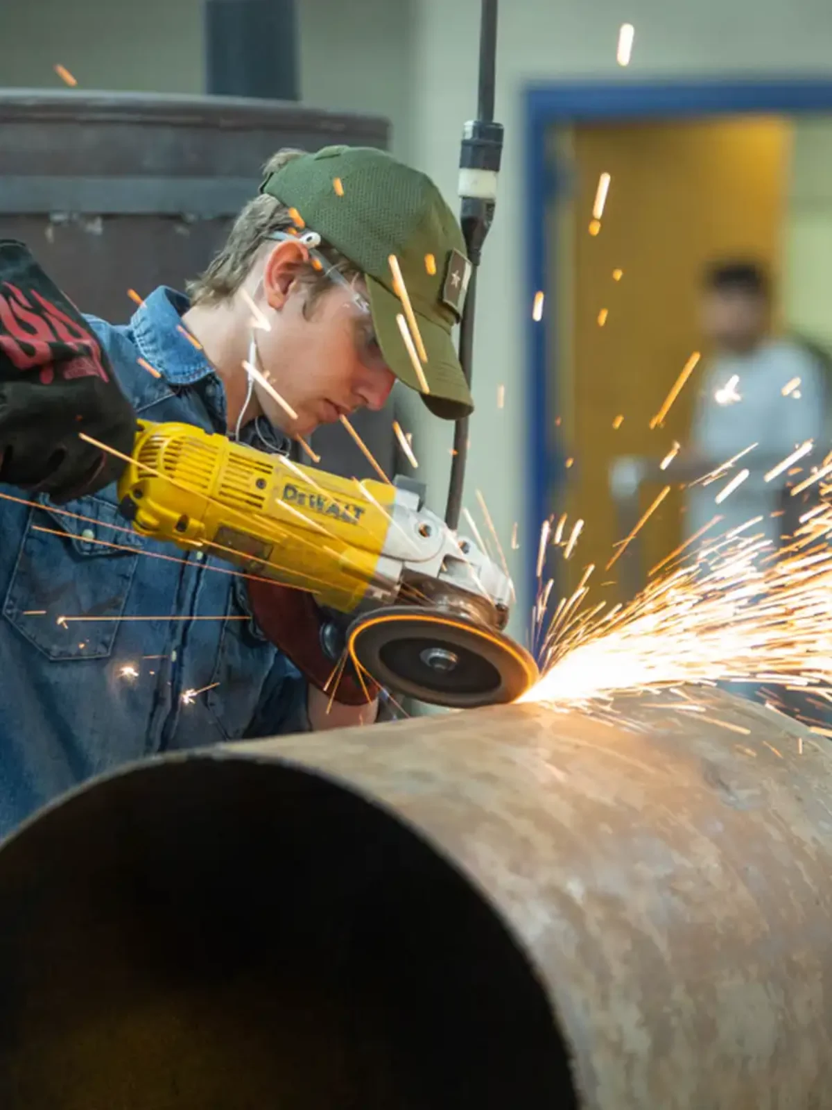 A young adult is in a workshop, using a saw to cut pipe. Sparks are flying from where he's cutting.