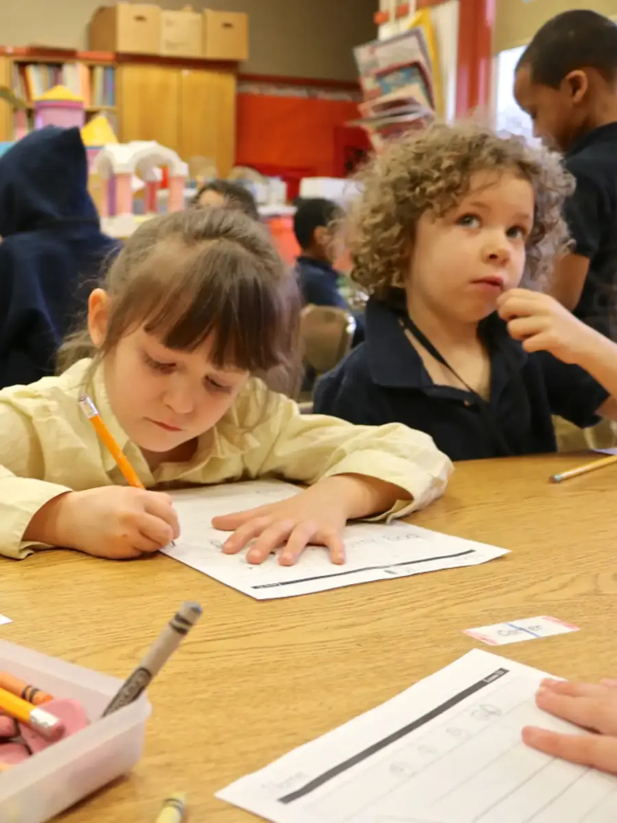 Two young children sit at a table. One is leaning over a worksheet with a pencil in her hand.