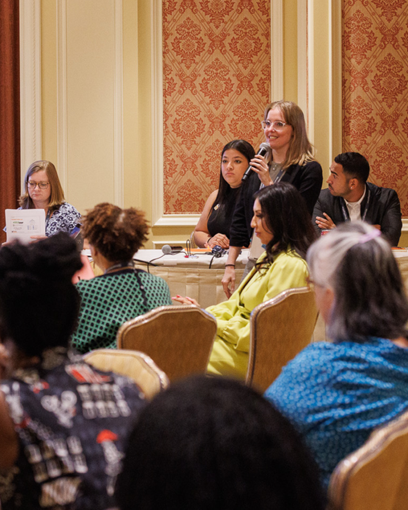 A woman stands with a microphone speaking in front of a conference workshop.
