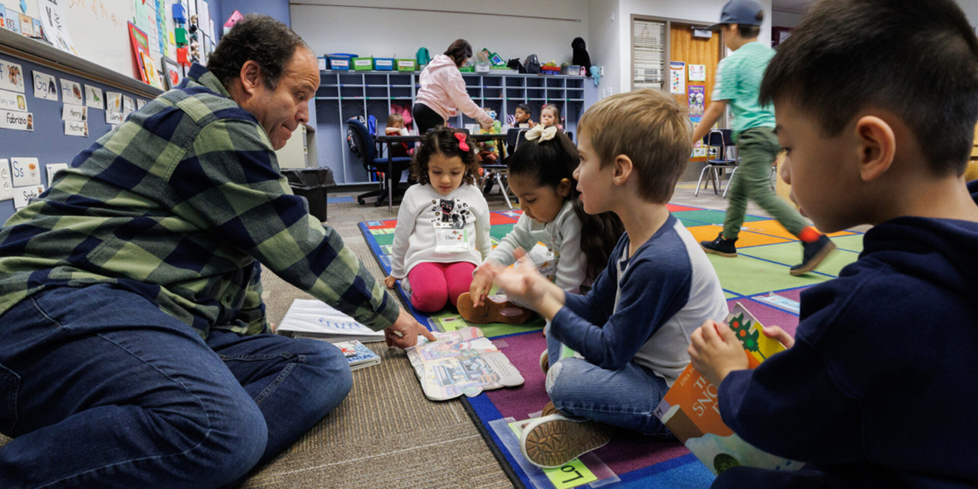 A teacher sits on the ground in front of three young children, pointing at a book laying open in front of them.