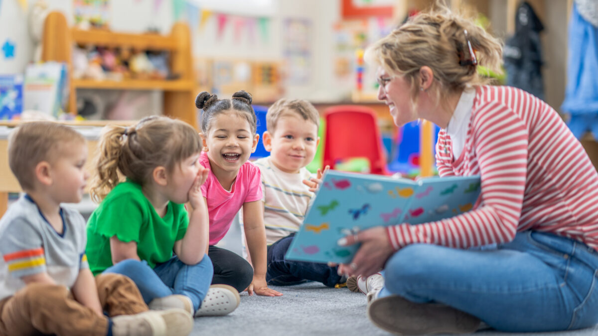 A group of young students sit on the ground in front of a teacher who's reading a book to them.