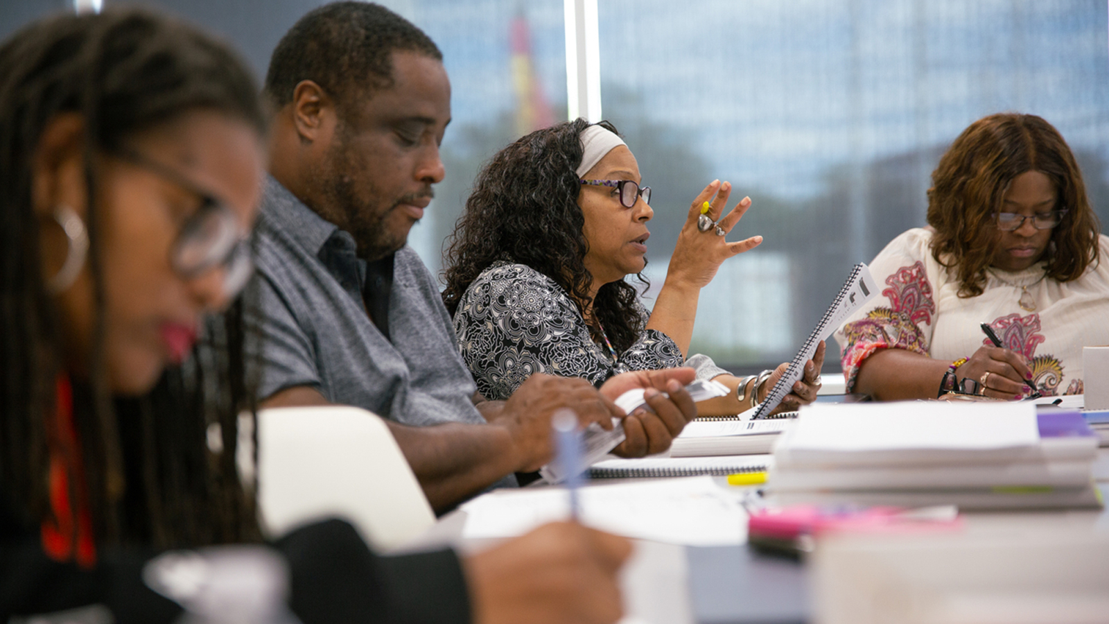A group of people sit at rows of tables. The camera is focused on one woman who's speaking as she looks at a booklet open in front of her.