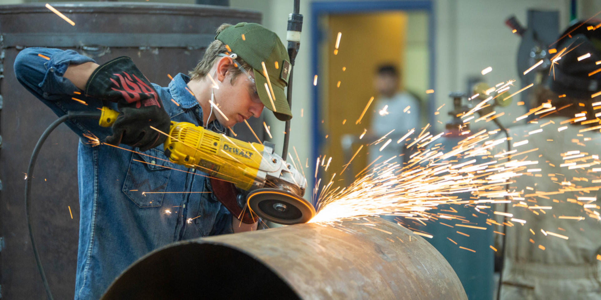 A young adult is in a workshop, using a saw to cut pipe. Sparks are flying from where he's cutting.