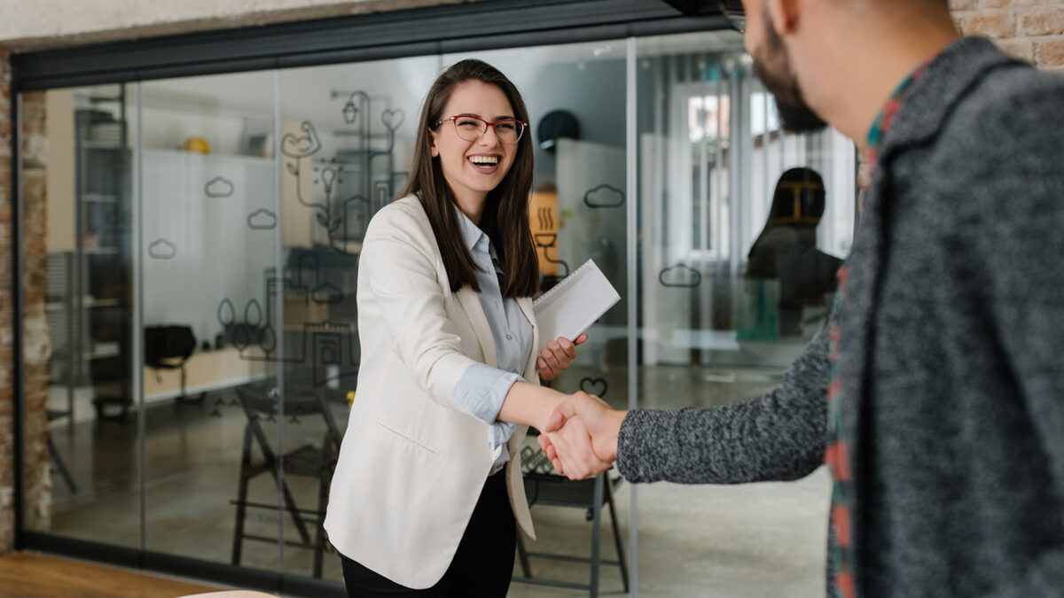 A young woman wearing a blazer shakes hands with someone as she walks in to a room.