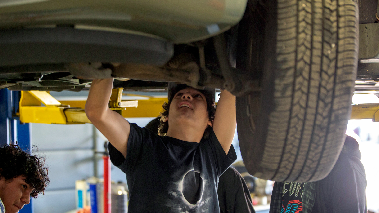 A young adult is working in a garage, standing underneath a car with his arms extended upward.