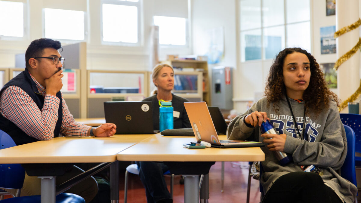 Three adults sit around a table, all looking in the same direction as if they're listening to someone outside of the photo speak.