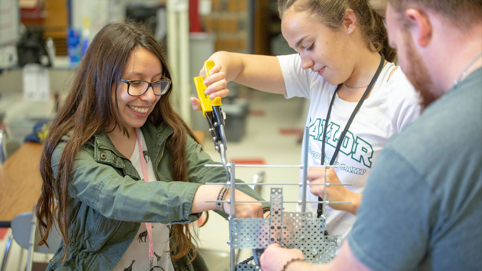 Two middle school-aged students are using pliers to hold together a device while an adult helps them hold it steady.