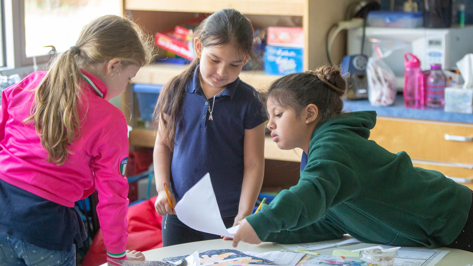 Three elementary aged children lean over a table together looking at a drawing.
