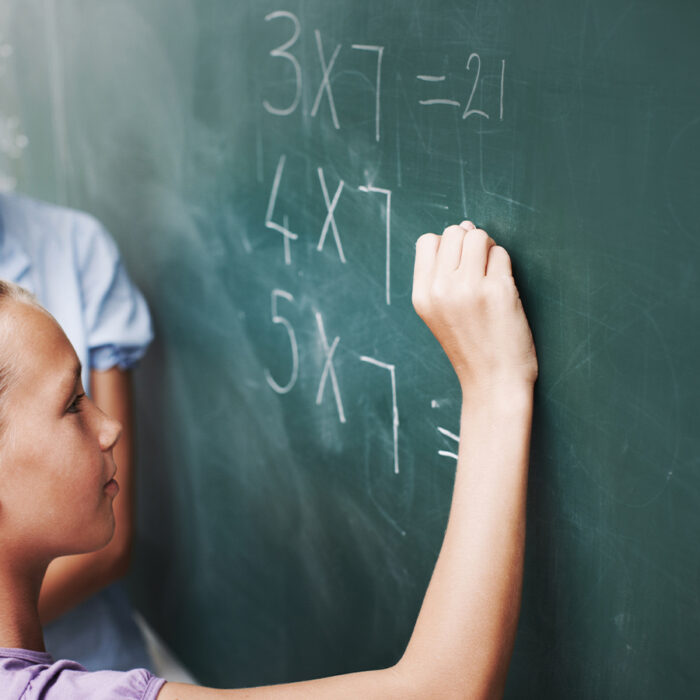 A young girl writes a math equation on a chalkboard while a teacher watches.