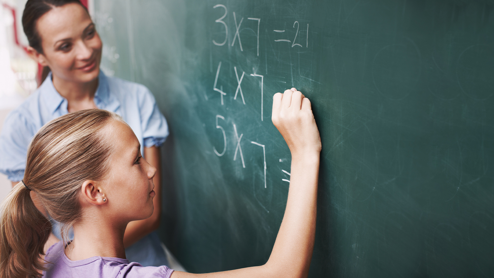 A young girl writes a math equation on a chalkboard while a teacher watches.