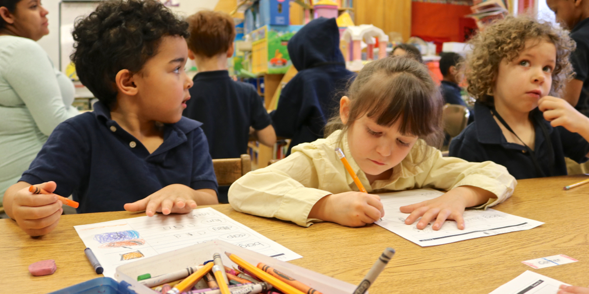 Three young children sit at a table. One is leaning over a worksheet with a pencil in her hand.