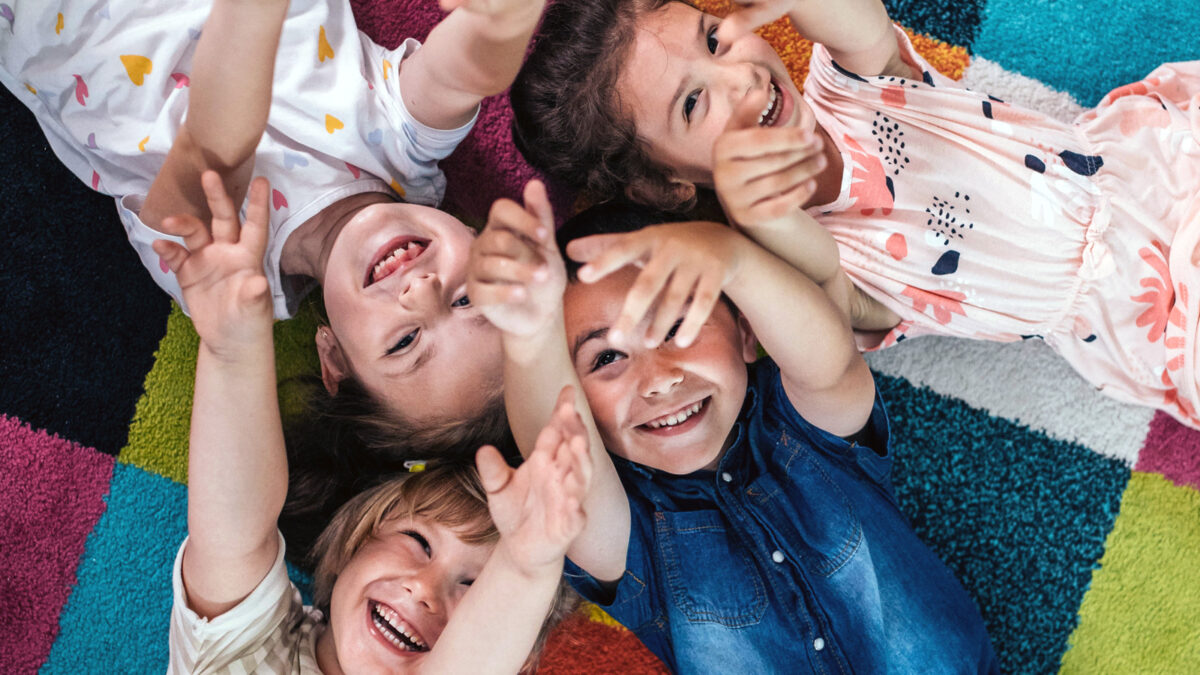 A group of kindergarten students lay on their backs on a rug, reaching up toward the camera and smiling