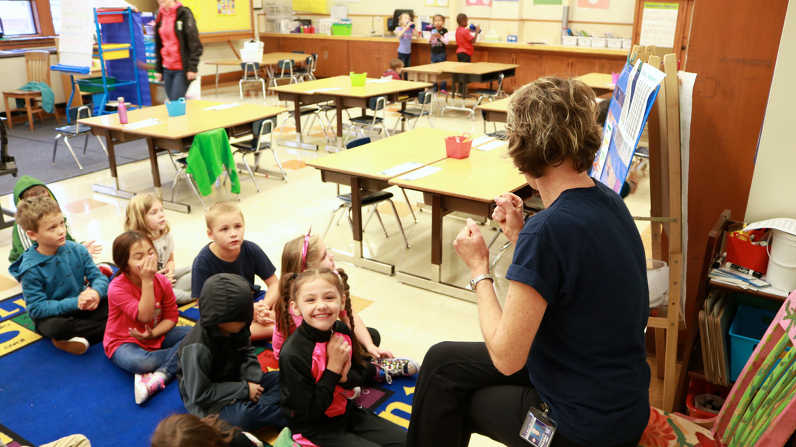 A teacher sits in front of a group of young students all sitting in a line. They're in an elementary school classroom.