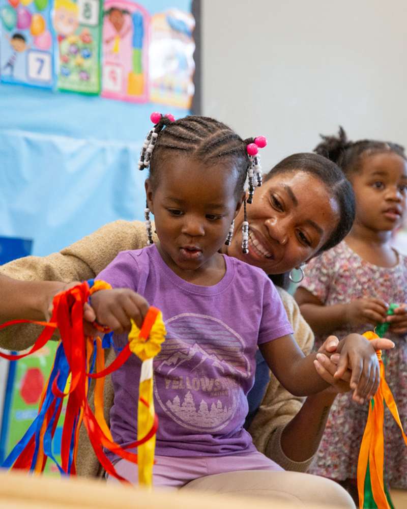 A kindergarten-aged student looks excitedly at ribbons that an adult is holding in front of her.