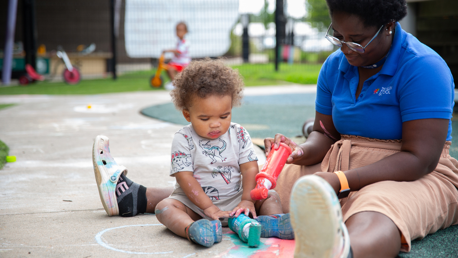 A toddler sits on the ground with an adult playing with rolling chalk.