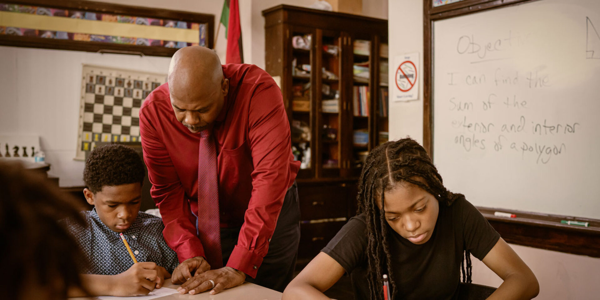 Two students sit at a desk. A teacher is leaning over to talk to one of them as they look at the student's work together.