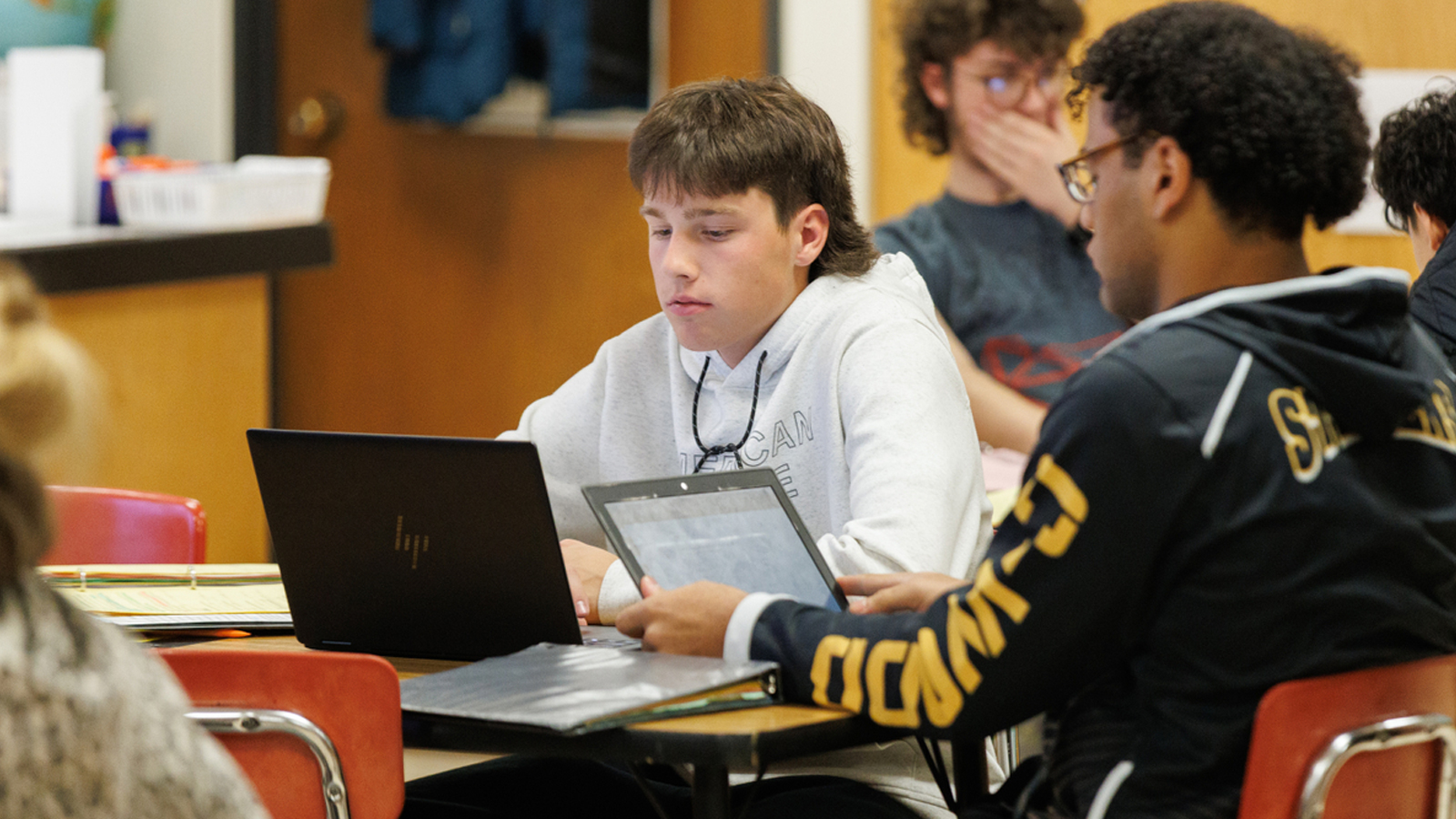 Two middle-school aged boys sit at a table working off of laptops.