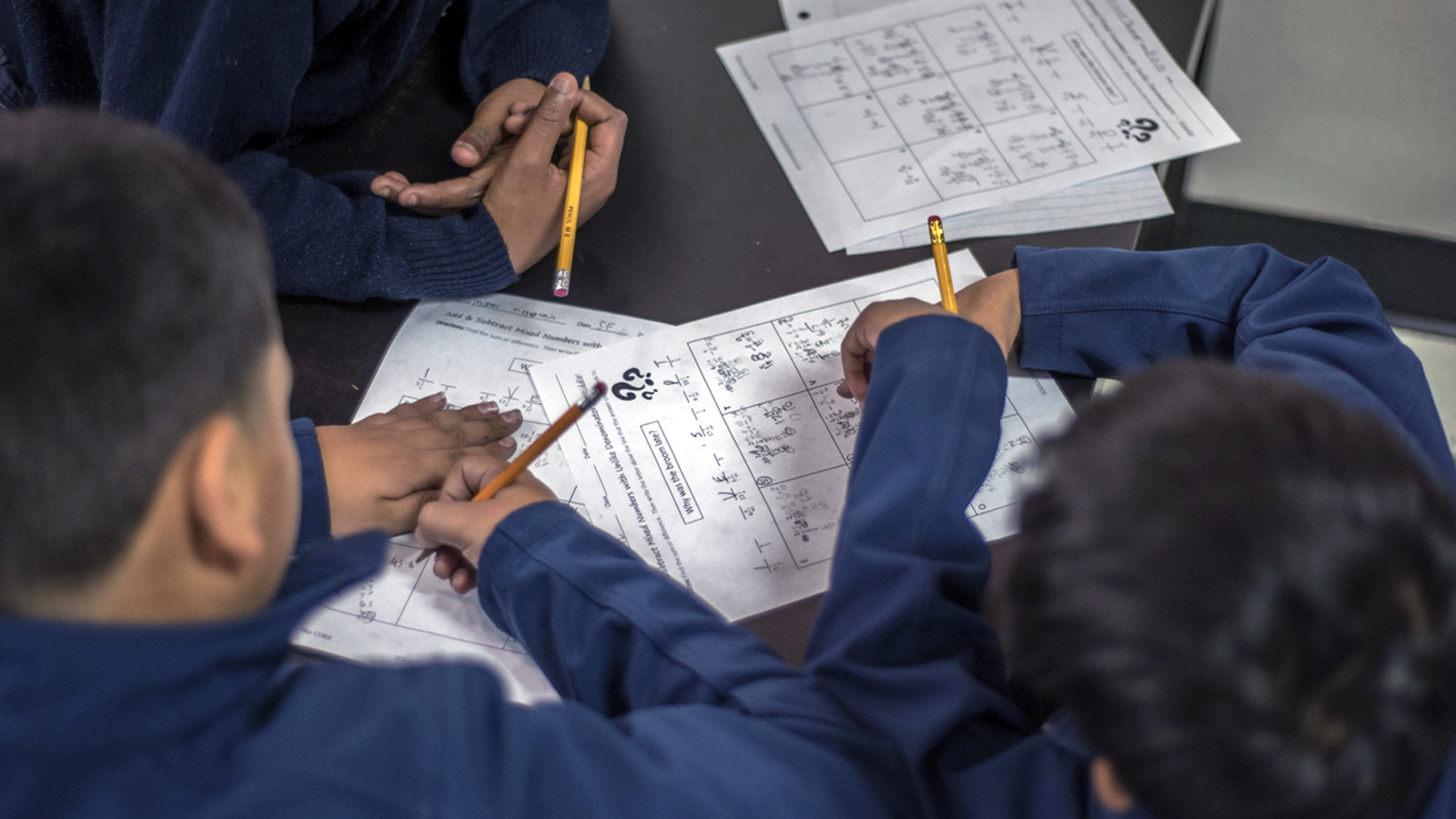 A close up shot of three students writing on worksheets. They appear to be learning division.