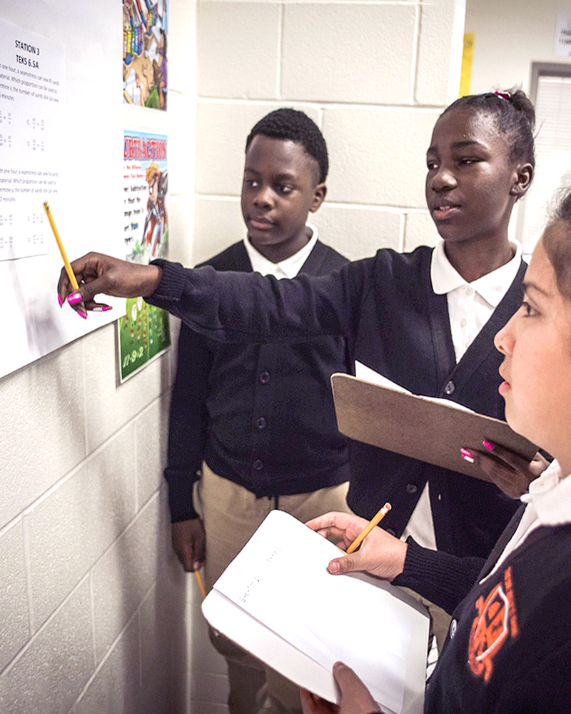 Three middle-school aged students are looking at a poster on the wall, taking notes on clipboards.