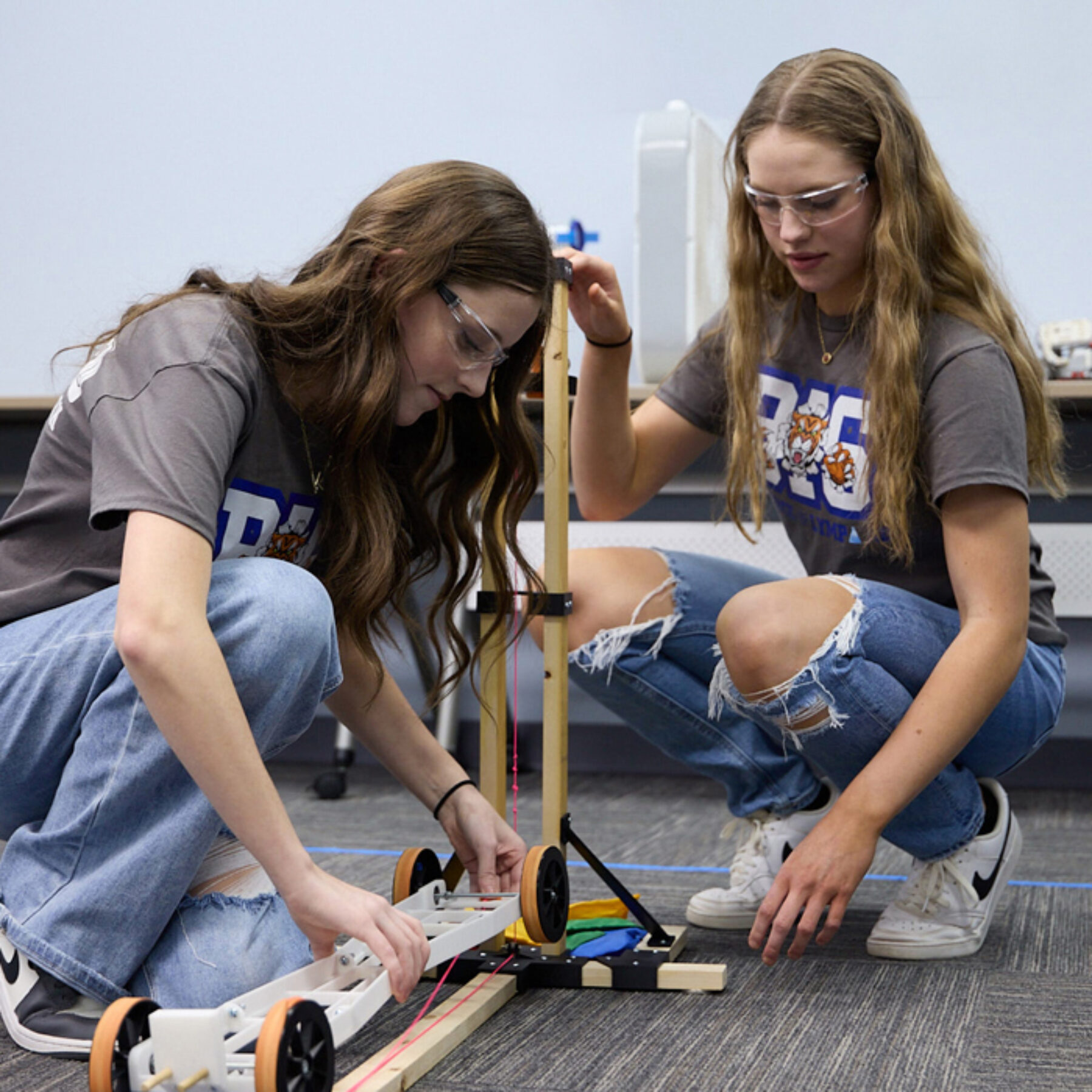 Two high-school aged girls kneel close to the ground as they build a device out of wood.