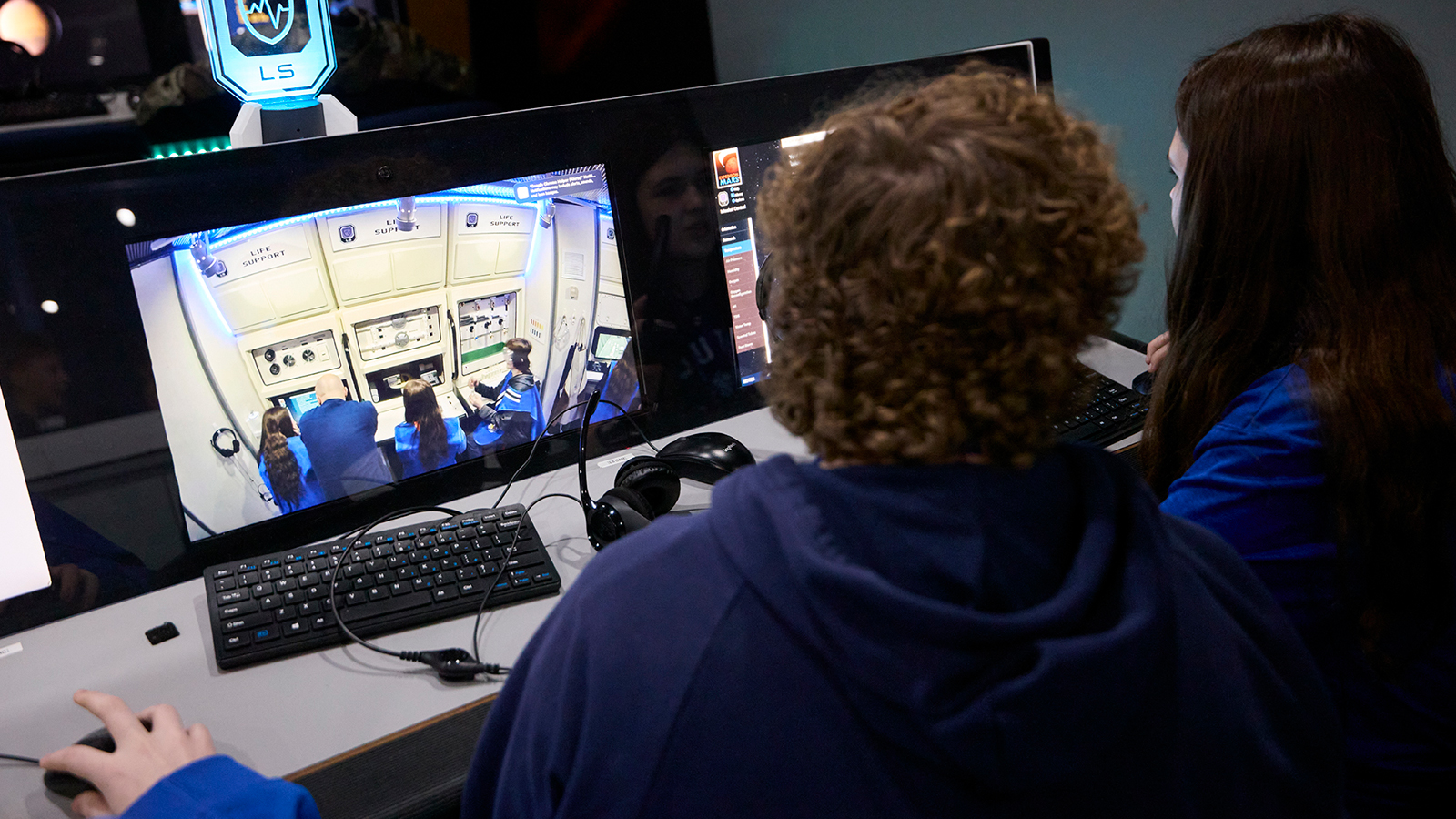 Two students look at a computer screen where they're watching their peers act out a space travel simulation.