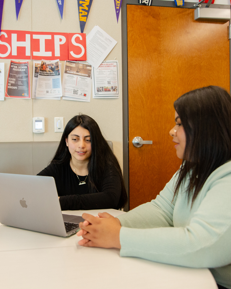 An academic advisor sits with a student at a table, looking at a laptop screen together.