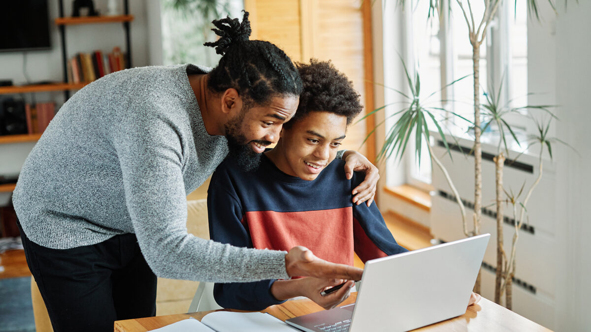 A parent and student look at a computer together. The parent seems to be congratulating his son on being accepted to a college.