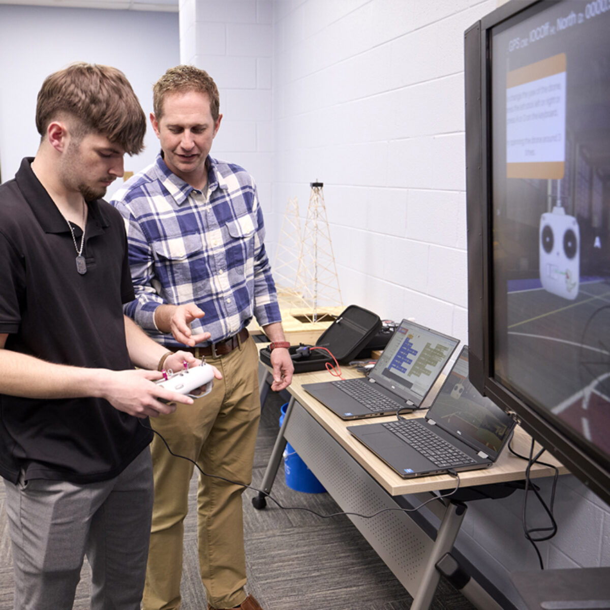 A student and teacher stand holding a controller as the student learns how to use a drone.