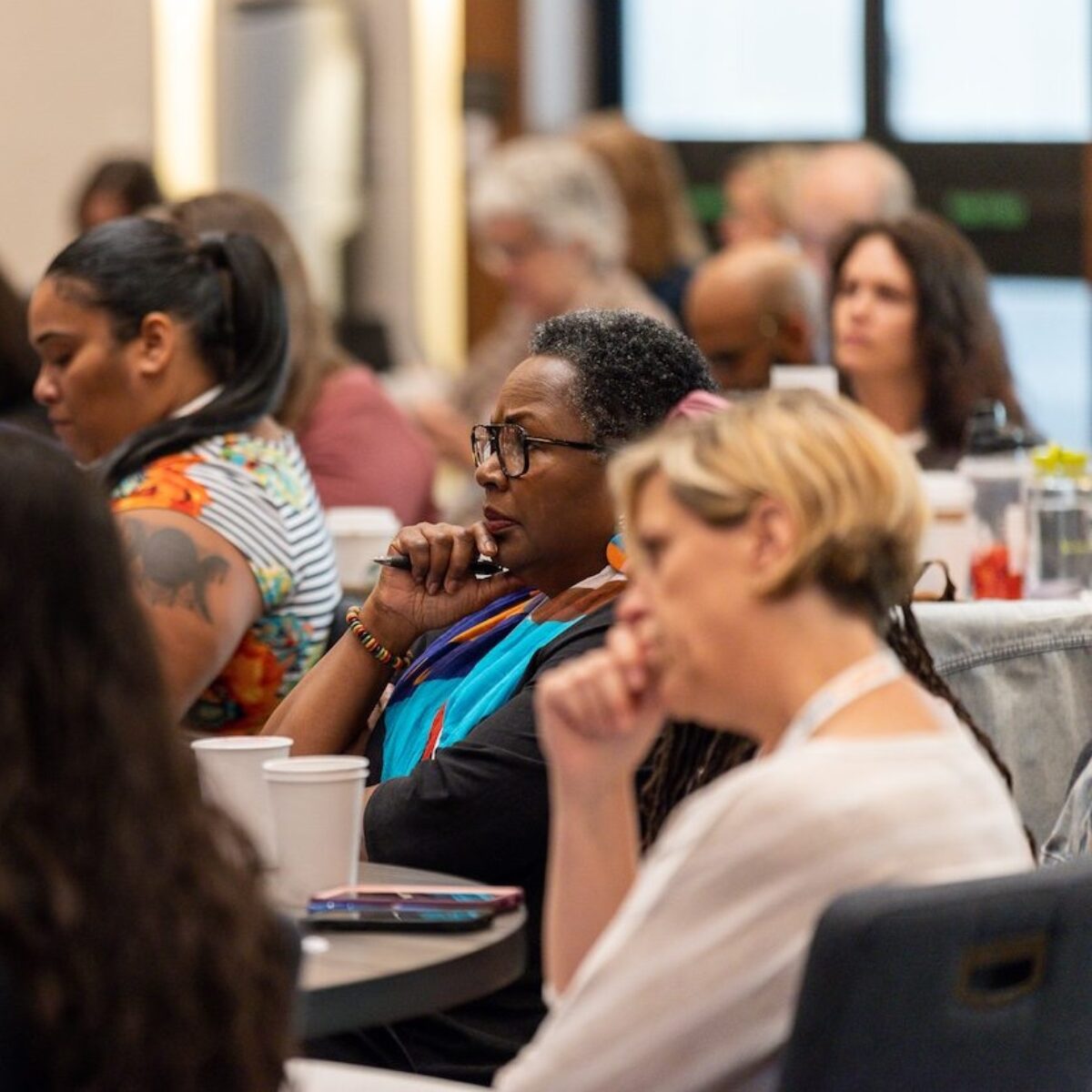 Two women sit at a table in a conference workshop room. They both have their hands on their chins, listening intently.