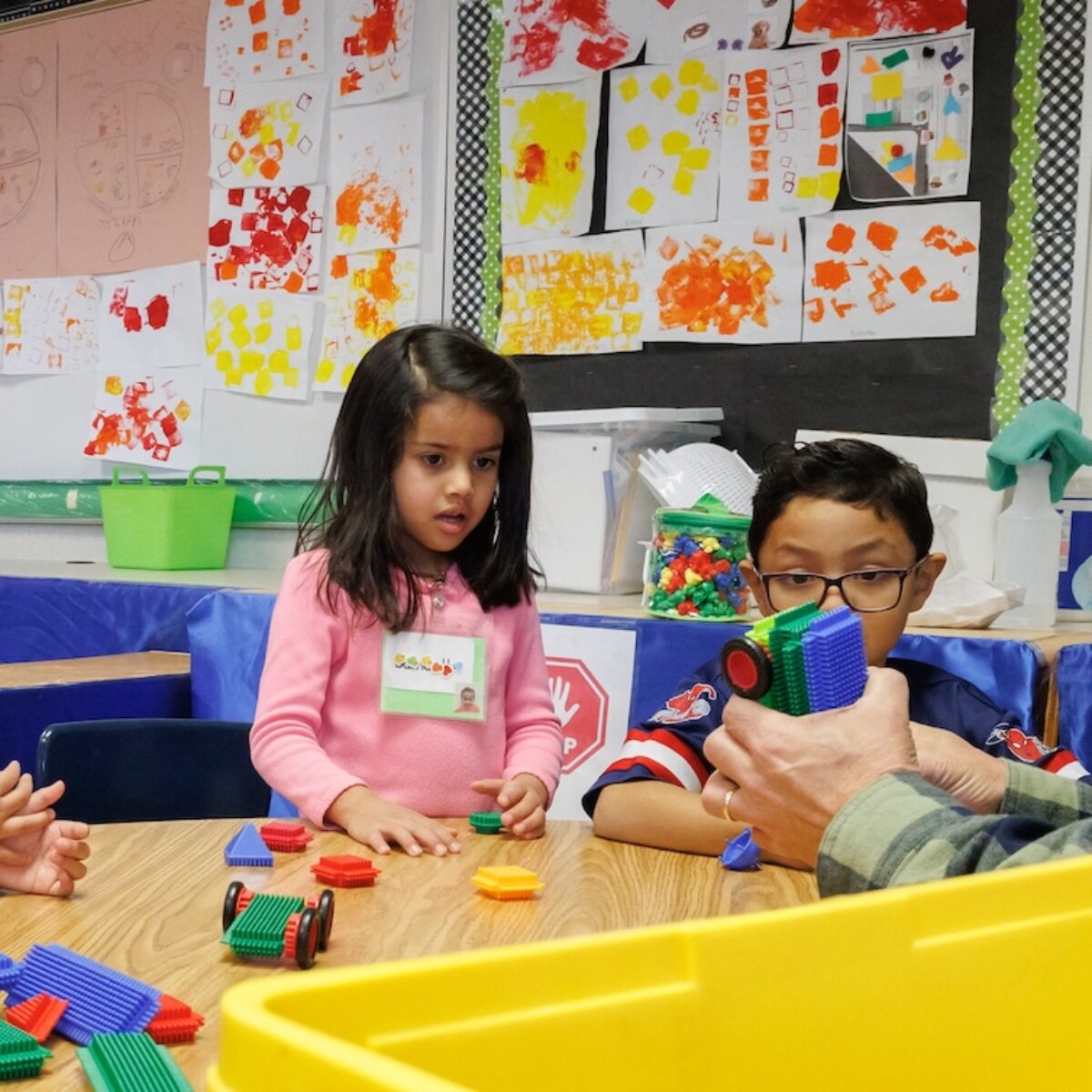 Three young learners from Salt Lake City play with blocks at a table. A teacher is building a car out of the blocks.