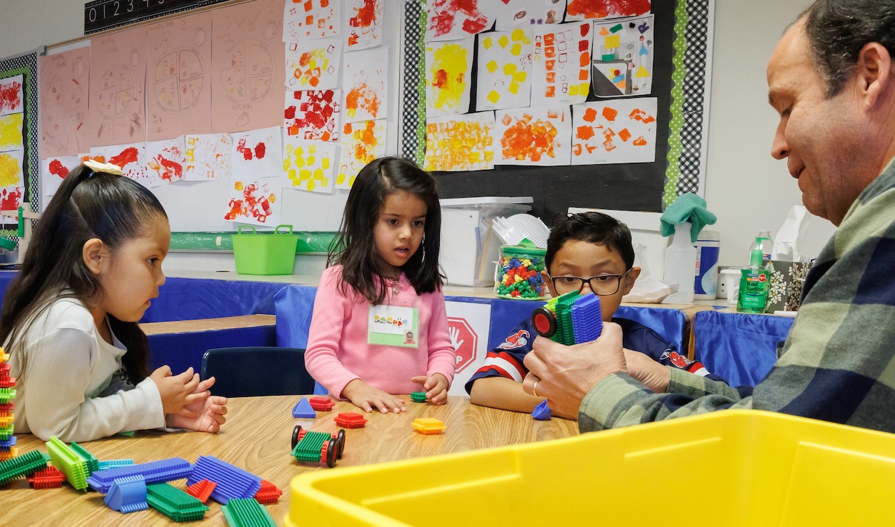 Three young learners from Salt Lake City play with blocks at a table. A teacher is building a car out of the blocks.